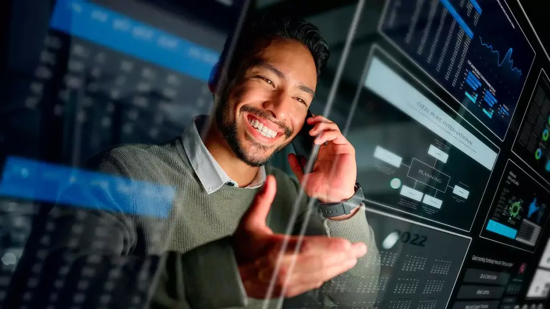 Homem sorrindo enquanto usa o celular diante de vários monitores